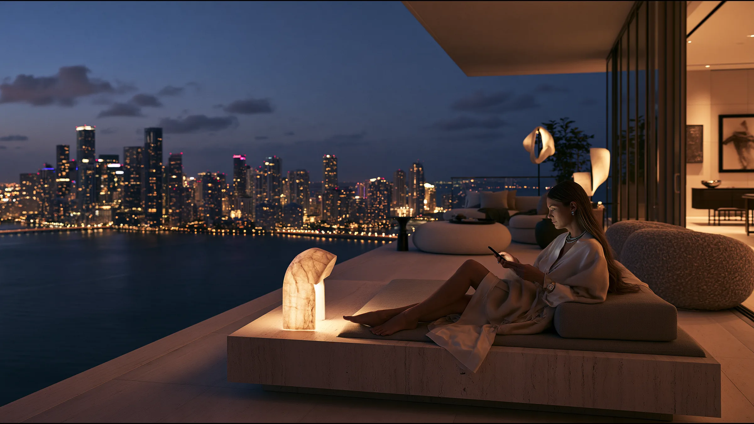 A woman on a vast penthouse terrace in Brickell at night, reading a reply on her phone with quiet relief, a travertine daybed and sculptural lamp beside her, Biscayne Bay and the lit Miami skyline spread out below