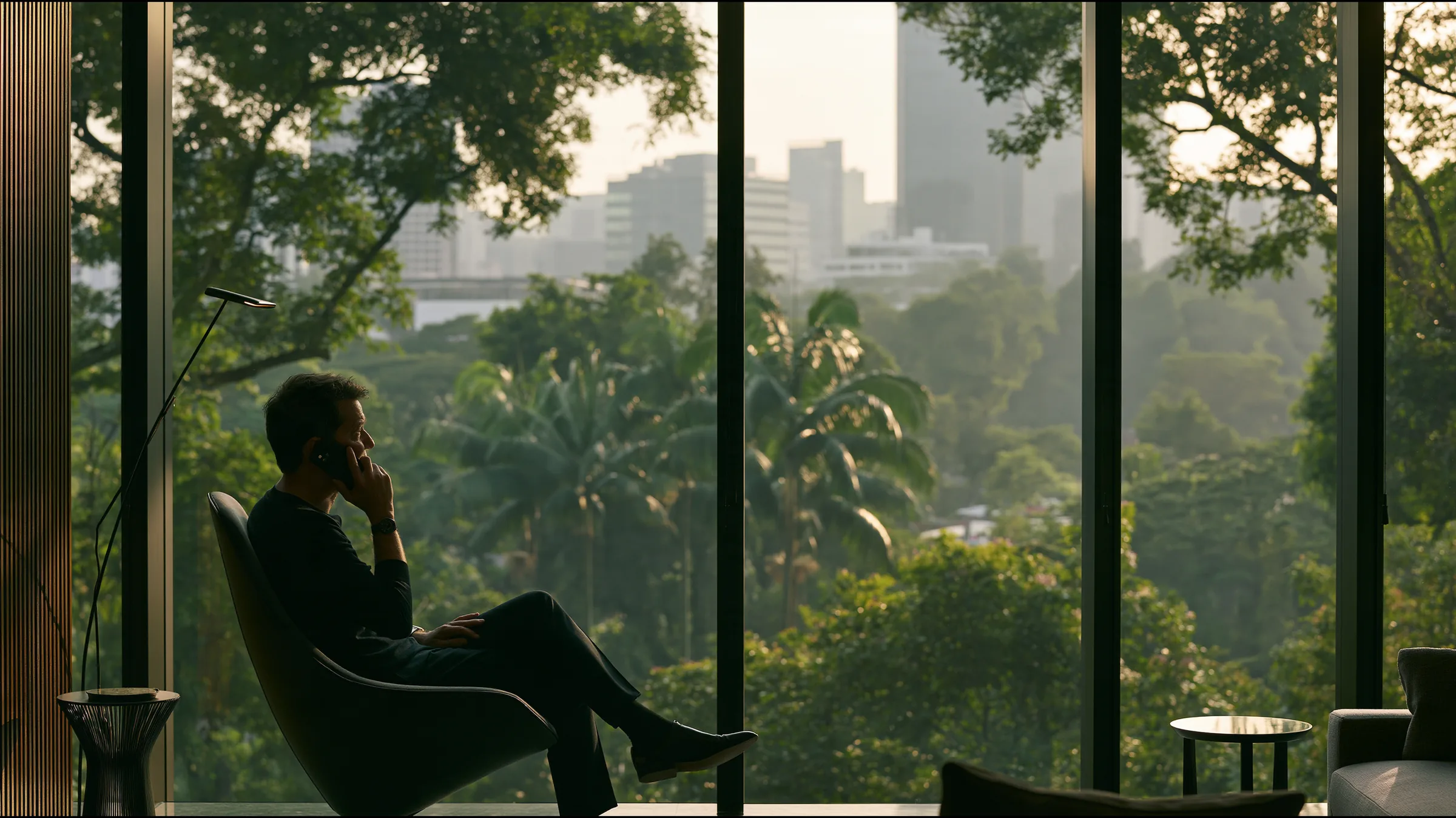 A medical director in his fifties in fine knit seated in a single sculptural chair in a vast calm consulting suite above Orchard Road, Singapore, on a phone call with gentle reassurance, tropical trees and towers beyond the glass