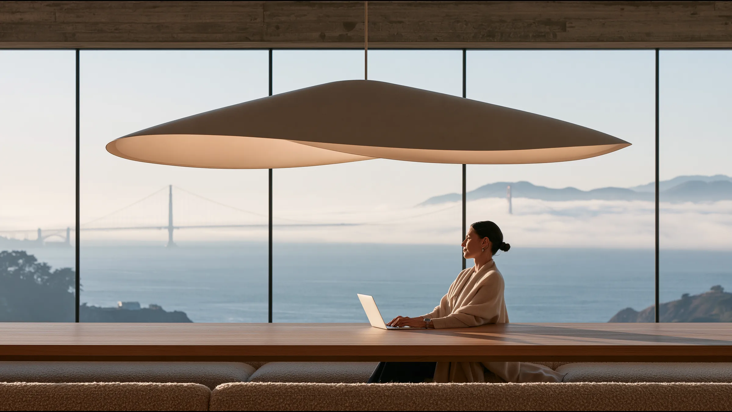 A physician in her forties alone at a vast oak table in a glass pavilion clinic in Pacific Heights, a single laptop open, a quiet look of delight, the Golden Gate Bridge and the bay in morning fog beyond the glass