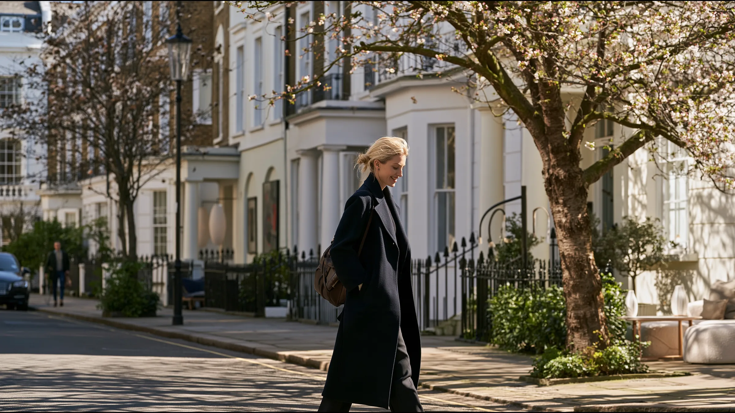 A locum GP in a navy wool coat walking along a wide sunlit Georgian terrace in Islington on a spring morning, smiling, a leather bag over her shoulder, blossom trees and white facades