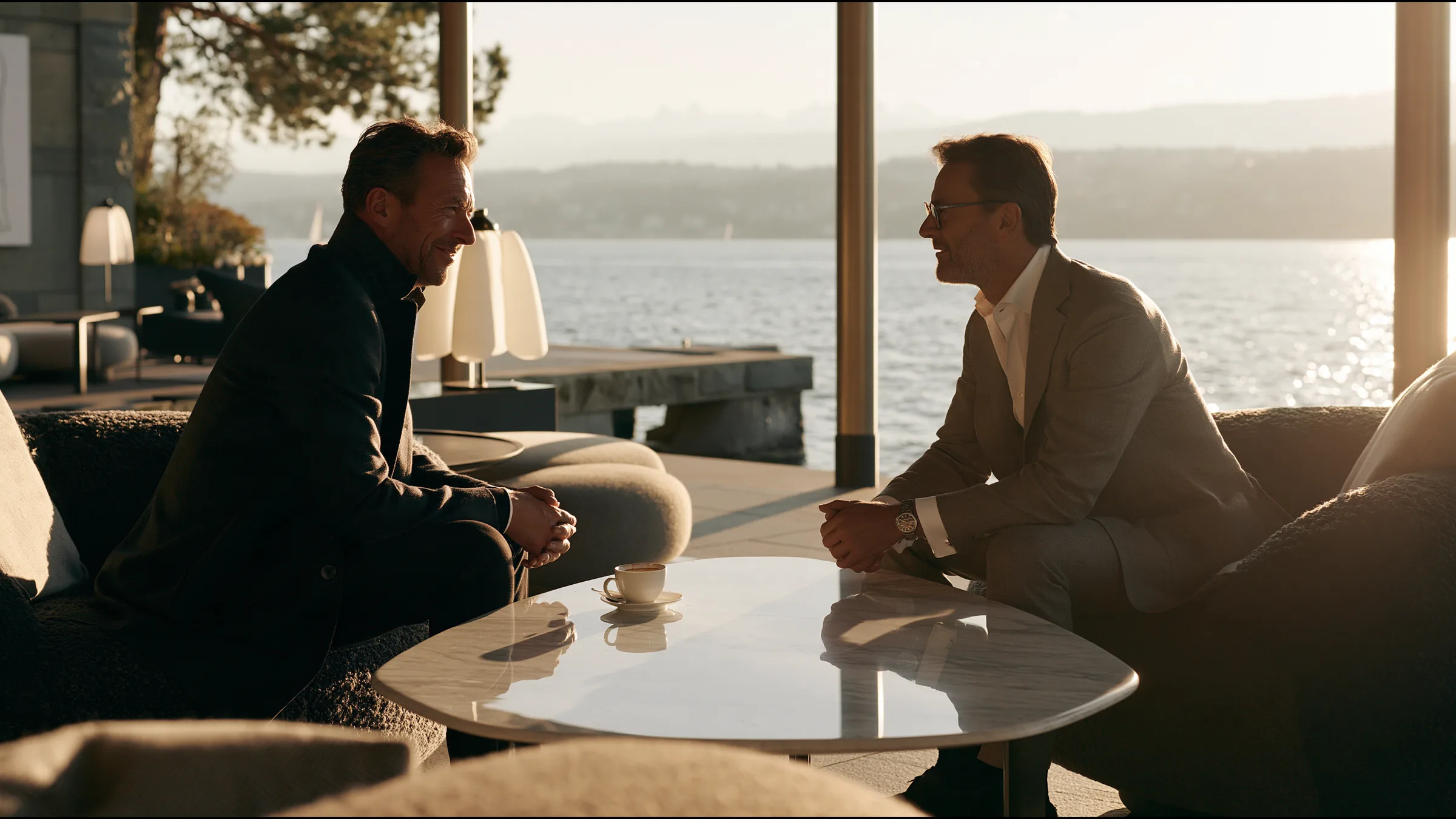 Two clinic partners in their forties in smiling over coffee at a marble table on a lakeside terrace in Seefeld, Zurich, the lake and the Alps in morning light