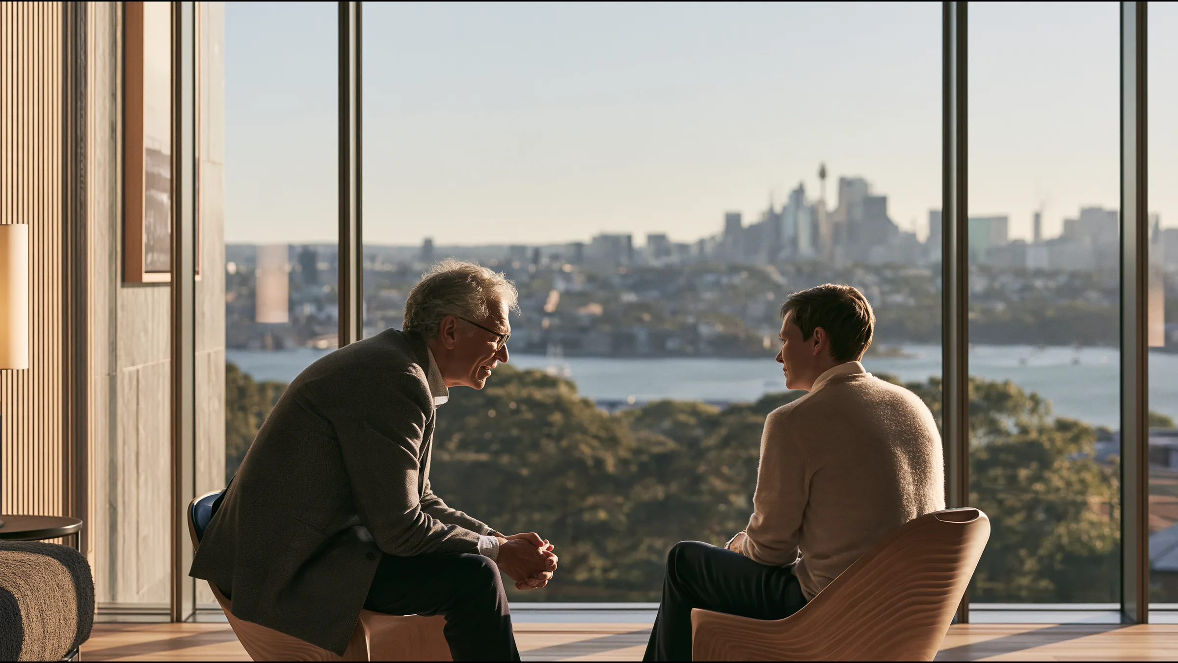A consultant physician in his sixties in an unstructured grey jacket leaning forward in conversation with a patient on two sculptural chairs in a vast calm Macquarie Street consulting suite in Sydney, the harbour beyond the glass