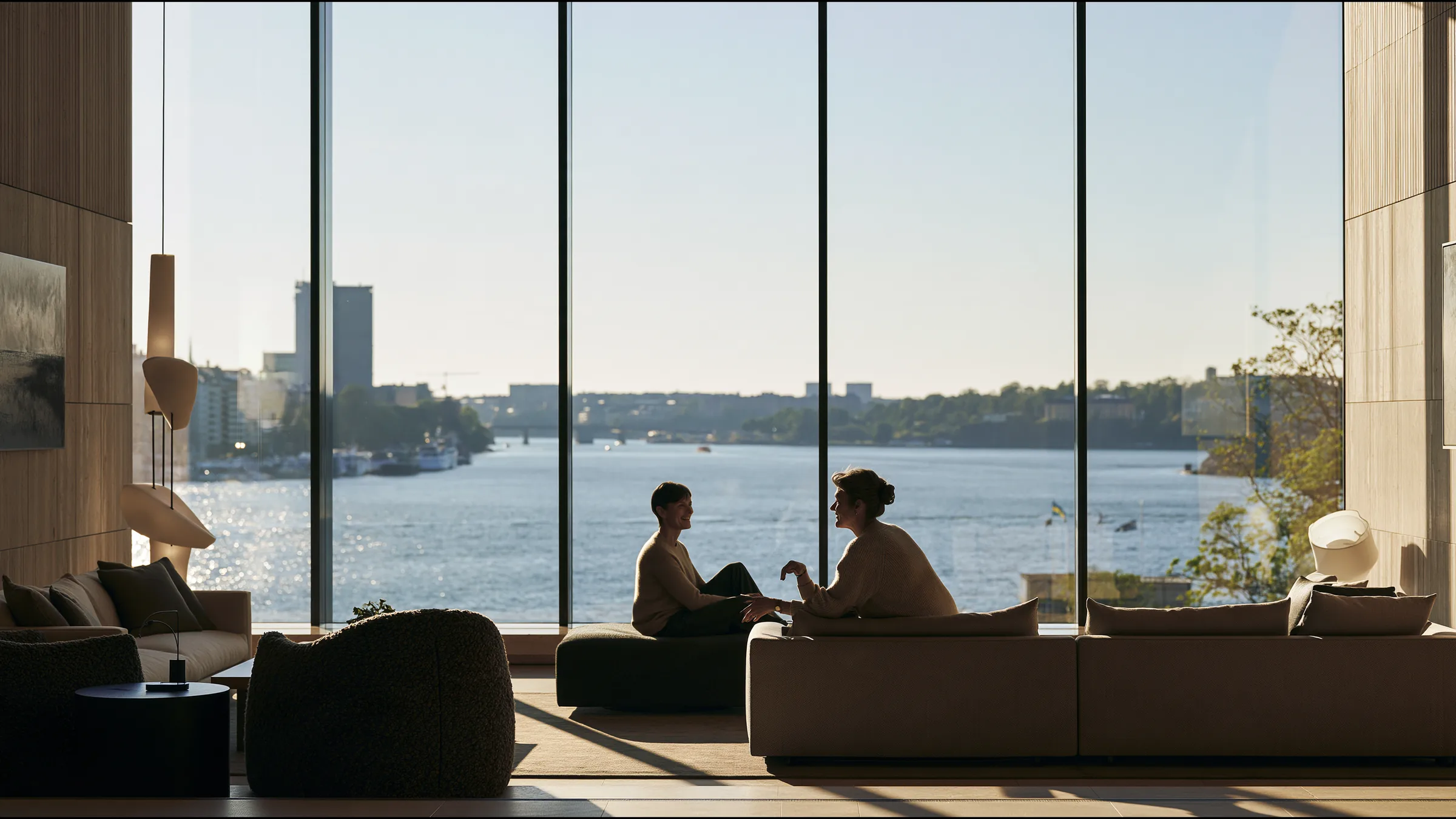 A nurse coordinator in her fifties in fine knit talking warmly with a returning patient by a vast tall window in a bright Östermalm clinic in Stockholm, the waterfront in clear northern light