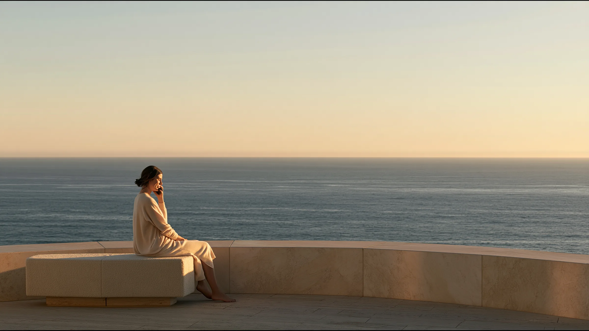 A woman in her thirties in fine athletic knitwear sitting on a stone bench on a clifftop above Bondi Beach after an early run, smiling as she talks to her doctor on the phone, the ocean vast at sunrise