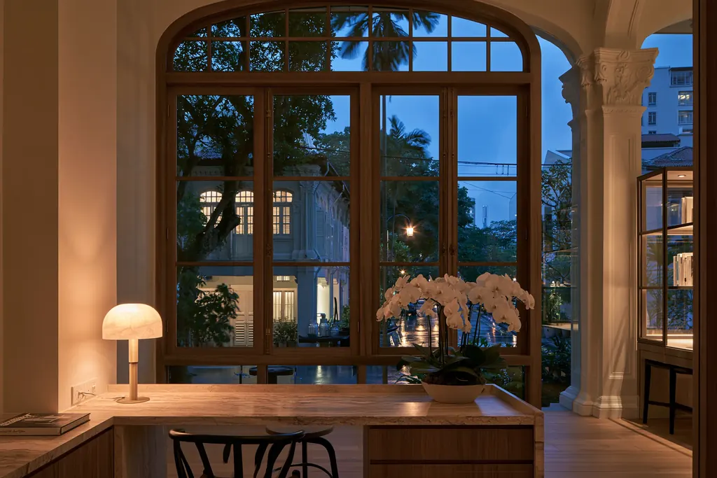 The front desk of a restored Singapore clinic at dusk, a lamp lit and tall timber windows onto the street